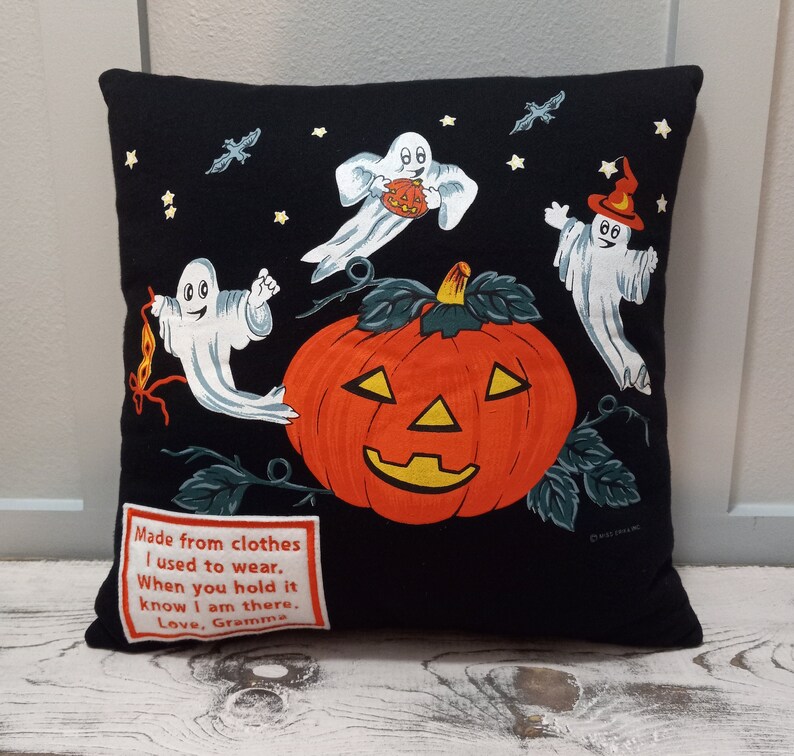 May include: Black Halloween-themed pillow featuring a large orange jack-o'-lantern with a smiling face, surrounded by three cartoon ghosts, bats, and stars. A patch with text reads "Made from clothes I used to wear. When you hold it know I am there. Love, Gramma."