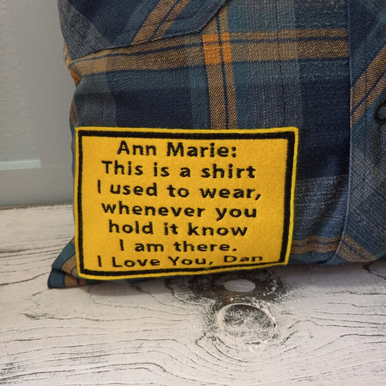 May include: A cushion made from a blue and gold plaid shirt. A yellow patch with black embroidered text reads: "Ann Marie: This is a shirt I used to wear, whenever you hold it know I am there. I Love You, Dan."