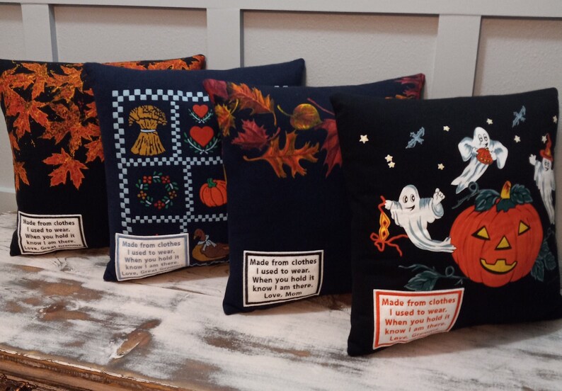 May include: Four decorative throw pillows with fall and Halloween themes. Designs include autumn leaves, a harvest scene, and ghosts with a jack-o'-lantern. Each pillow has a black background and a sewn-on label with text.