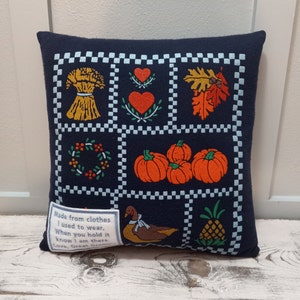May include: Navy blue decorative pillow with a patchwork design featuring fall-themed images like pumpkins, leaves, and a wheat bundle. The pillow includes a small tag with text.