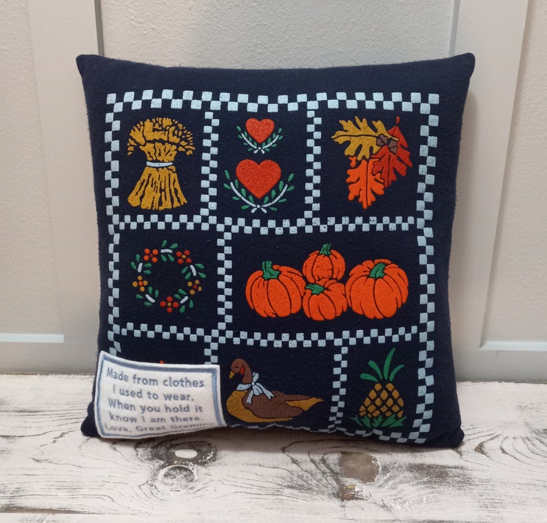 May include: Navy blue decorative pillow with a patchwork design featuring fall-themed images like pumpkins, leaves, and a wheat bundle. The pillow includes a small tag with text.