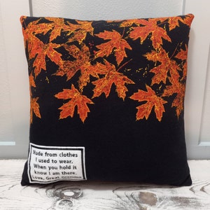 May include: Square black pillow with a pattern of orange and yellow maple leaves. The pillow has a white label with the text: "Made from clothes I used to wear. When you hold it know I am there. Love, Great Gramma."