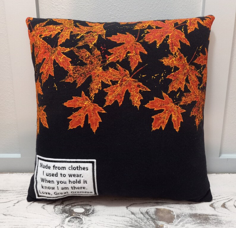 May include: Square black pillow with a pattern of orange and yellow maple leaves. The pillow has a white label with the text: "Made from clothes I used to wear. When you hold it know I am there. Love, Great Gramma."