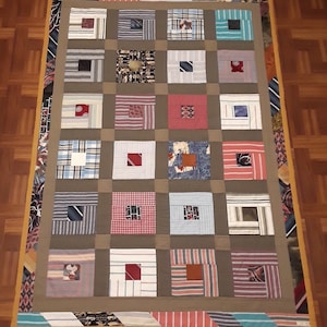 May include: A handmade quilt with a patchwork design. The quilt features squares of various patterned fabrics, including stripes and plaids, framed by a brown border. The edges are finished with a decorative border of ties and a yellow trim.