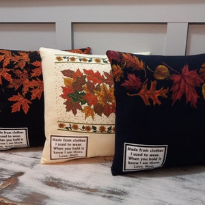 May include: Three decorative throw pillows with fall leaf designs. One pillow is black with orange leaves, another is cream with a leaf cluster, and the third is navy with red leaves. Each pillow has a white label with text.