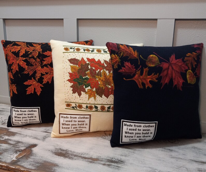May include: Three decorative throw pillows with fall leaf designs. One pillow is black with orange leaves, another is cream with a leaf cluster, and the third is navy with red leaves. Each pillow has a white label with text.