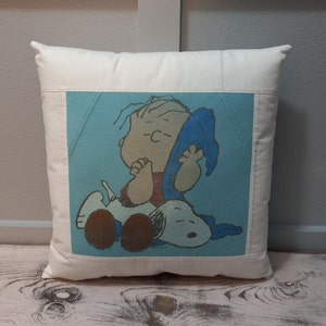 May include: Square off-white pillow with a light blue center panel featuring a cartoon image of Linus and Snoopy. Linus is wearing a red shirt and Snoopy is white with brown spots. The pillow has a simple, classic design.