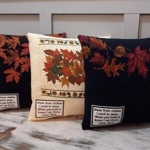 May include: Three decorative throw pillows with fall leaf designs. One pillow is black with orange leaves, another is cream-colored with a mix of fall leaves, and the third is navy blue with red and orange leaves. Each pillow has a white label with text.