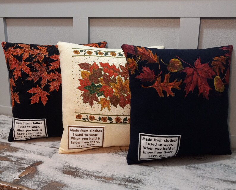 May include: Three decorative throw pillows with fall leaf designs. One pillow is black with orange leaves, another is cream-colored with a mix of fall leaves, and the third is navy blue with red and orange leaves. Each pillow has a white label with text.