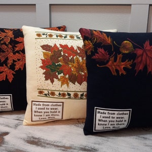 Puede incluir: Tres cojines decorativos con diseños de hojas de otoño. Uno es negro con hojas naranjas, otro es de color crema con un arreglo central de hojas, y el tercero es azul marino con hojas rojas y naranjas. Cada cojín tiene una pequeña etiqueta de texto.