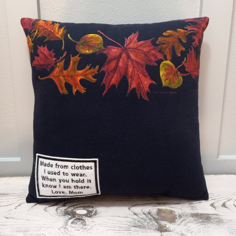 May include: A navy blue square pillow with a border of colorful autumn leaves. The pillow has a white patch with the text "Made from clothes I used to wear. When you hold it know I am there. Love, Mom."