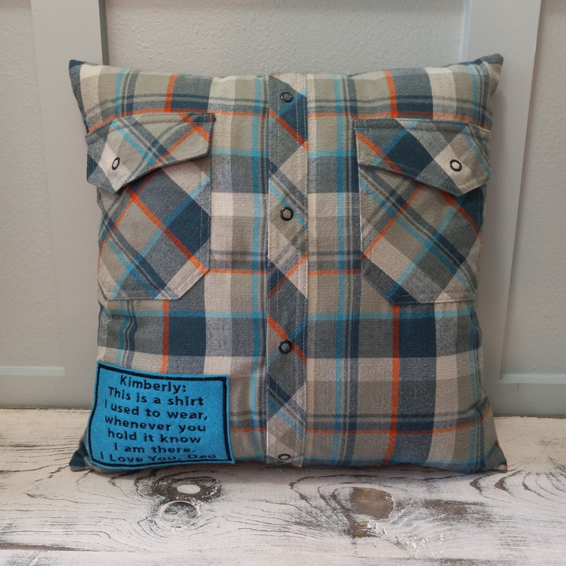 May include: A square cushion crafted from a plaid shirt, in shades of blue, grey, and orange. It has two shirt pockets and a blue patch with the text: "Kimberly: This is a shirt I used to wear... I Love You, Dad."