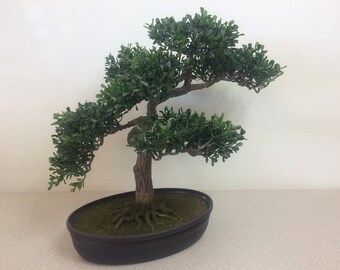 May include: A small, artificial bonsai tree with green leaves and a brown trunk. The tree is planted in a dark brown, oval-shaped pot. The tree's branches extend outward, creating a natural, miniature tree appearance.