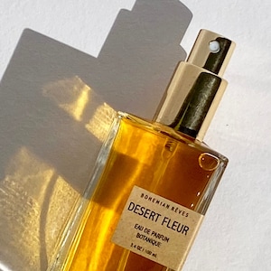 Close-up of Bohemian Rêves Desert Fleur Perfume Mist bottle casting deep red shadows, reflecting the warm, earthy essence of patchouli and vanilla. A bold, sun-drenched fragrance inspired by desert heat and golden light.