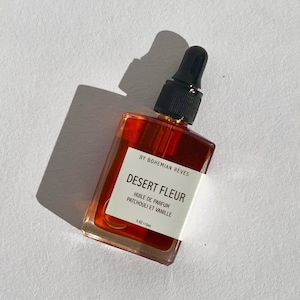 May include: A clear glass bottle with a black dropper top filled with a red liquid. The bottle has a white label with the text "BY BOHEMIAN RÊVES DESERT FLEUR HUILE DE PARFUM PATCHOULI ET VANILLE 1.5 OZ/115ml".