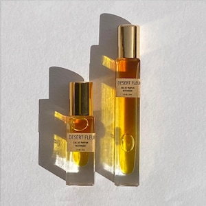 May include: Two glass bottles of perfume with gold caps and labels that read "Desert Fleur Eau de Parfum Botanique". The bottles are on a white surface.