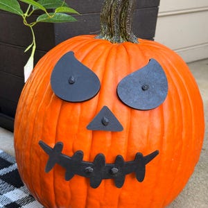 May include: A large, round orange pumpkin with a carved face for Halloween. The face is made of black metal cutouts for the eyes, nose, and mouth. The pumpkin sits on a black and white checkered mat.