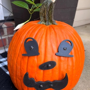 May include: A large, bright orange pumpkin with a carved face for Halloween. The face is made of black metal cutouts for the eyes, nose, and mouth. The pumpkin has a green stem and is sitting on a black and white checkered surface.