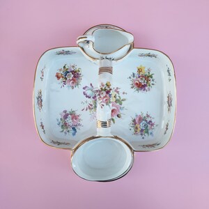 Pode incluir: Um prato de servir de porcelana branca com uma borda dourada e um padrão floral. O prato tem uma alça embutida e duas tigelas menores, uma de cada lado.
