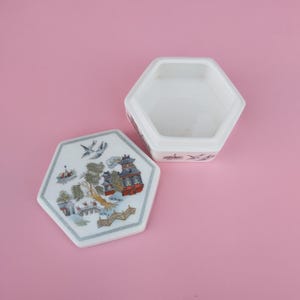 May include: A white hexagonal box with a lid. The lid features a colourful design of a Chinese-style building, trees, and birds. The box is empty.