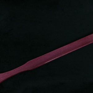 May include: A long, wooden paddle with a rounded end. The paddle is a deep purple color and has a smooth, polished finish.