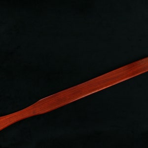 May include: A long, flat wooden paddle with a rounded handle. The paddle is made of a dark reddish-brown wood and has a smooth, polished finish.