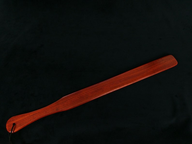 May include: A long, flat wooden paddle with a rounded handle. The paddle is made of a dark reddish-brown wood and has a smooth, polished finish.
