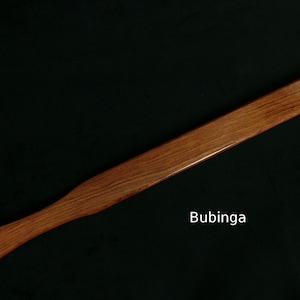 May include: A long, wooden paddle made of bubinga wood. The paddle is smooth and has a rich reddish-brown color.