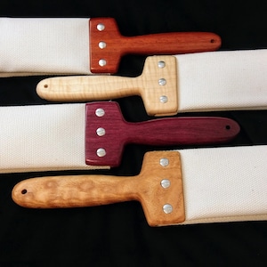May include: Four leather strop tools with wooden handles. The strops are white canvas and the handles are made of different types of wood. The handles are red, natural wood, purple, and brown.
