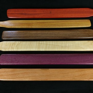 May include: Six wooden paddles of different wood types and colors. The paddles are arranged in a row from top to bottom, with the top paddle being red, the second paddle being light brown, the third paddle being dark brown, the fourth paddle being light wood, the fifth paddle being purple, and the sixth paddle being light orange.