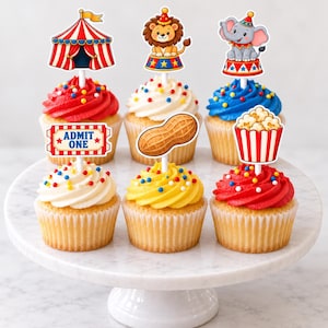Op de afbeelding: Zes cupcakes met circus-thema decoraties, waaronder een rood-wit gestreepte tent, een leeuw, een olifant, een pinda, een popcornemmer en een rood ticketbordje met de tekst ADMIT ONE. De cupcakes zijn voorzien van rode, witte, gele en blauwe glazuur.