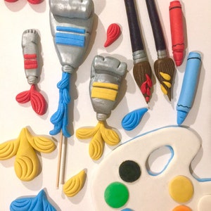May include: An assortment of art-themed decorations, including silver paint tubes with blue and yellow accents, paintbrushes, crayons, and a white artist's palette with colorful paint circles. The decorations are in shades of red, blue, yellow, and green.