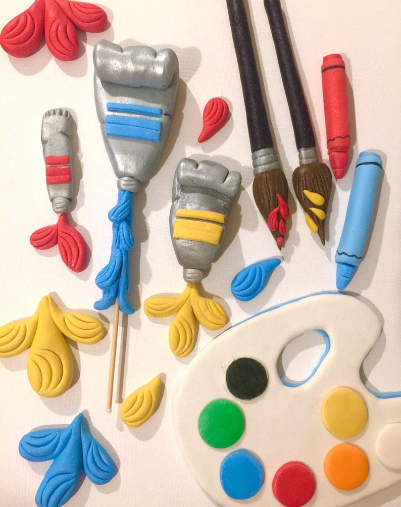 May include: An assortment of art-themed decorations, including silver paint tubes with blue and yellow accents, paintbrushes, crayons, and a white artist's palette with colorful paint circles. The decorations are in shades of red, blue, yellow, and green.