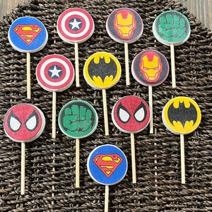 May include: A collection of superhero-themed cupcake toppers. The toppers feature iconic symbols like Superman's logo, Captain America's shield, Iron Man's mask, the Hulk's fist, Spider-Man's mask, and the Batman logo. Each topper is round and attached to a wooden stick.