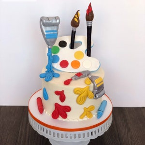 May include: A two-tiered cake decorated with art-themed elements. The cake features a white base with colorful accents, including paintbrushes, a palette, paint tubes, and crayons. The cake sits on a white and orange cake stand.