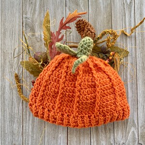 May include: A crocheted pumpkin hat in orange with green leaves on top. The hat is sitting on a wooden background.
