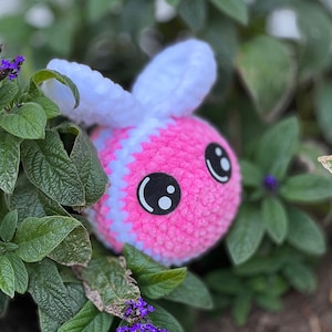 May include: A pink and white crocheted bee plush toy with black eyes. The bee is sitting in a bed of green leaves.
