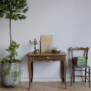 May include: A vintage wooden desk with a single drawer, a decorative candelabra, and a vintage poster. A small bouquet of flowers sits on the desk. A wooden chair holds a stack of books. A large potted tree is on the left.