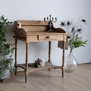 May include: A light brown wooden console table featuring a single central drawer with a small knob, a raised back shelf, and four turned legs. A lower shelf connects the legs, and a metal candelabra sits on the top surface of the wooden table.