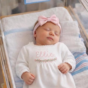 May include: A newborn baby wearing a white long-sleeved dress with the name "Ellie" embroidered in pink, along with a floral design. The baby also wears a pink bow headband. The baby is lying in a hospital bassinet.