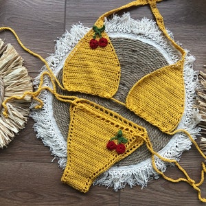 May include: Yellow crochet bikini top and bottom with red cherry embellishments. The bikini is laid out on a white fringed surface.