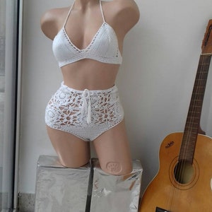 K&ouml;nnte beinhalten: Wei&szlig;er Bikini aus H&auml;kelarbeit mit Oberteil und hoch sitzender Hose. Das Oberteil hat einen Halterneck-Stil mit V-Ausschnitt. Die Hose hat einen hohen Bund und weite Beine.
