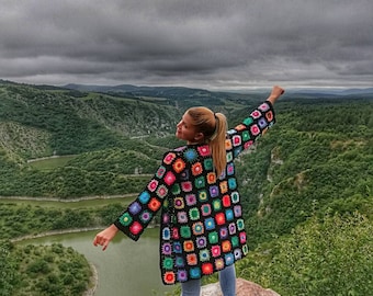 May include: A woman wearing a colorful crochet cardigan stands on a rocky cliff overlooking a winding river. The sky is overcast with dark clouds.