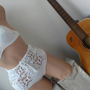 K&ouml;nnte beinhalten: Wei&szlig;er Bikini-Oberteil und hoch taillierte Hose aus H&auml;kelarbeit. Das Oberteil ist ein Halstuch mit Dreiecksform. Die Hose hat einen hohen Bund und ein spitzendes Muster.