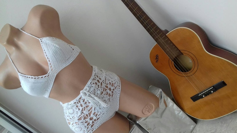 K&ouml;nnte beinhalten: Wei&szlig;er Bikini-Oberteil und hoch taillierte Hose aus H&auml;kelarbeit. Das Oberteil ist ein Halstuch mit Dreiecksform. Die Hose hat einen hohen Bund und ein spitzendes Muster.