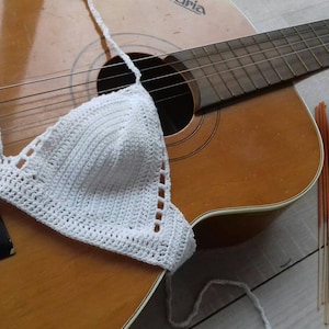 K&ouml;nnte beinhalten: Wei&szlig;es H&auml;kel-Bikini-Oberteil mit dreieckiger Form, das auf einer braunen Akustikgitarre mit hellbraunem Finish liegt. Die Gitarre hat ein Schallloch und Saiten. Es gibt auch mehrere orangefarbene R&auml;ucherst&auml;bchen auf einer hellbraunen Oberfl&auml;che.