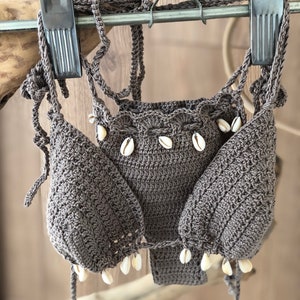 May include: A brown crocheted bikini top with white shell accents. The top is hanging on a wooden hanger.