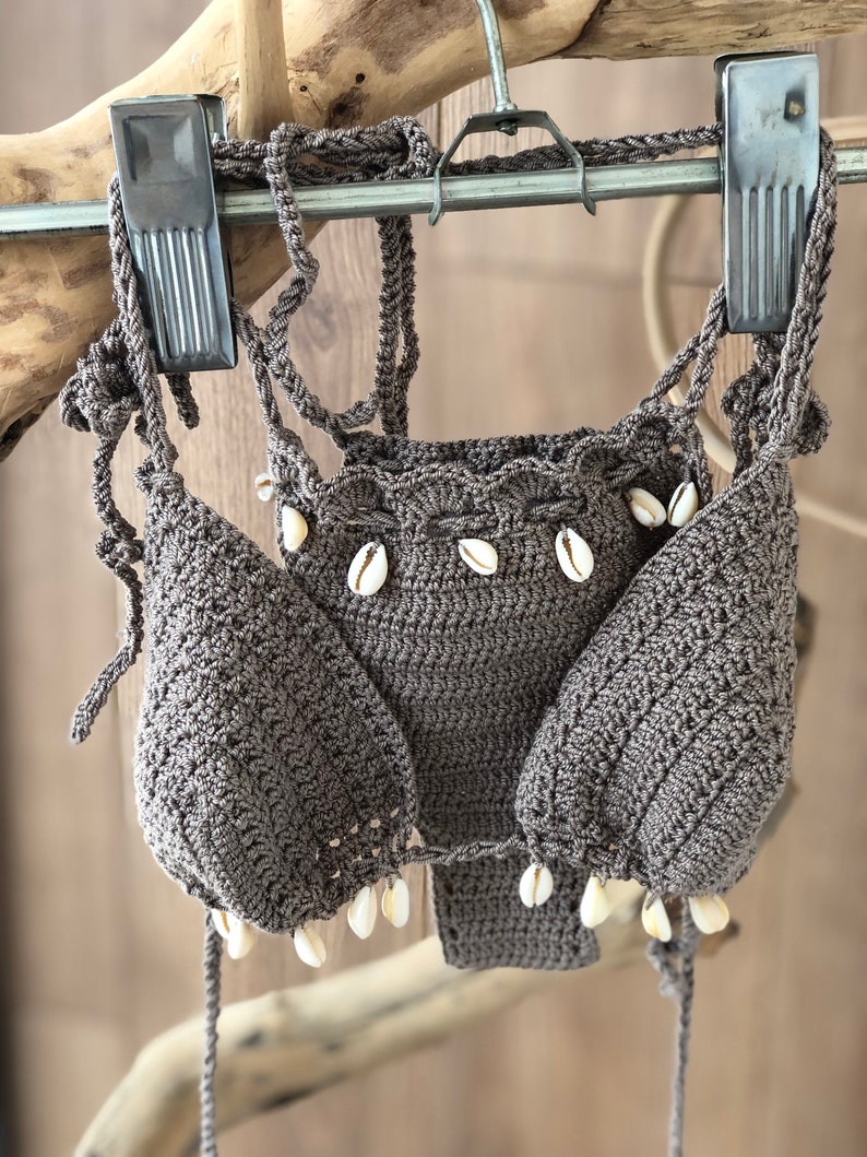 May include: A brown crocheted bikini top with white shell accents. The top is hanging on a wooden hanger.
