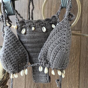 May include: A brown crocheted bikini top with white shell accents. The top is hanging on a wooden hanger.