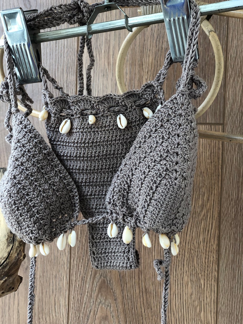 May include: A brown crocheted bikini top with white shell accents. The top is hanging on a wooden hanger.